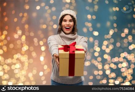 christmas, holidays and people concept - happy smiling young woman in knitted winter hat and sweater holding gift box over festive lights background. young woman in knitted winter hat holding gift box