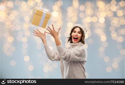 christmas, holidays and people concept - happy smiling young woman in knitted winter hat and sweater catching gift box over festive lights background. young woman in winter hat catching gift box
