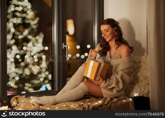 christmas, holidays and people concept - happy smiling young woman in pullover sitting on windowsill with gift box at home at night. happy woman with christmas gift at home at night