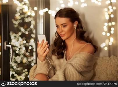 christmas, holidays and people concept - happy smiling young woman in pullover with smartphone and earphones listening to music sitting at window at home. woman with smartphone and earphones on christmas