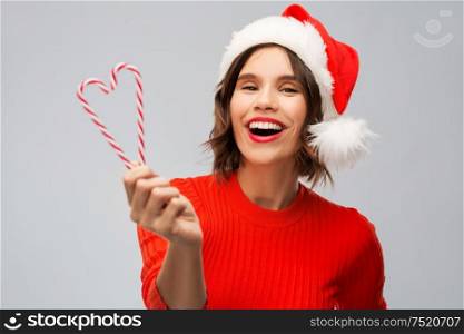 christmas, holidays and people concept - happy smiling young woman in santa helper hat with candy canes over grey background. happy young woman in santa hat on christmas