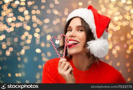 christmas, holidays and people concept - happy smiling young woman in santa helper hat licking candy canes over grey background. woman in santa hat licks candy canes on christmas