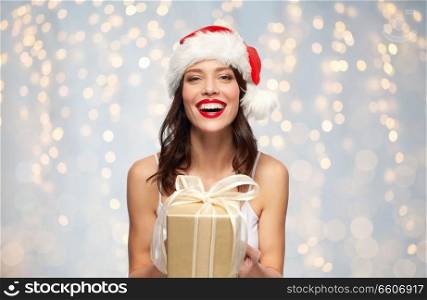 christmas, holidays and people concept - happy smiling young woman with red lipstick in santa hat holding present over festive lights background. woman in santa hat with christmas gift