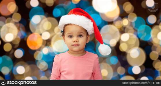 christmas, holidays and people concept - little baby girl in santa hat over lights background. little baby girl in santa hat at christmas