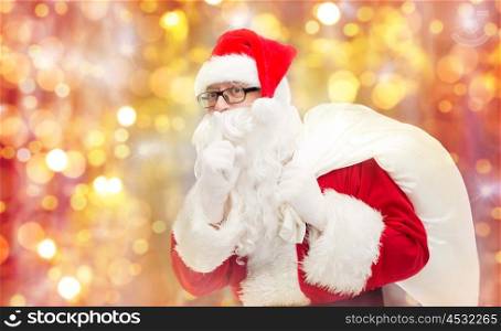 christmas, holidays and people concept - man in costume of santa claus with bag making hush gesture over lights background