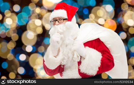 christmas, holidays and people concept - man in costume of santa claus with gifts bag making hush gesture over lights background. santa claus with gifts bag over christmas lights