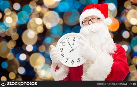 christmas, holidays and people concept - man in costume of santa claus with clock pointing finger to twelve over lights background. santa claus with twelve on clock at christmas