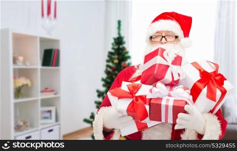 christmas, holidays and people concept - man in costume of santa claus with gift boxes over home room background. santa claus with christmas gifts at home