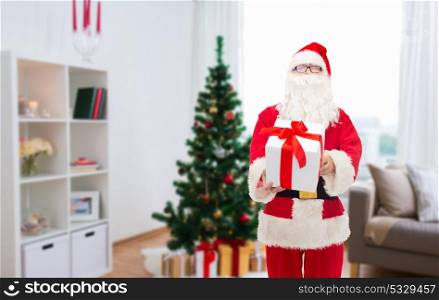 christmas, holidays and people concept - man in costume of santa claus with gift box over home room background. santa claus with christmas gift at home