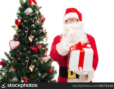 christmas, holidays and people concept - man in costume of santa claus with gift box and tree making hush gesture