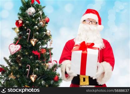 christmas, holidays and people concept - man in costume of santa claus with gift box and tree over blue lights background