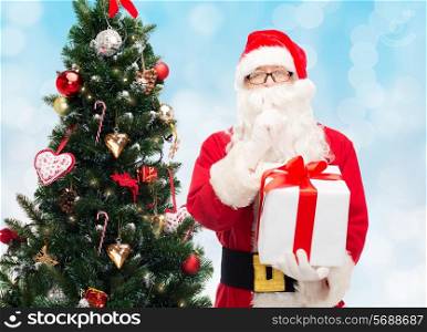 christmas, holidays and people concept - man in costume of santa claus with gift box and tree making hush gesture over blue lights background