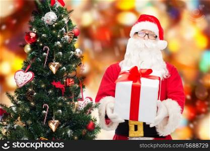 christmas, holidays and people concept - man in costume of santa claus with gift box and tree over red lights background