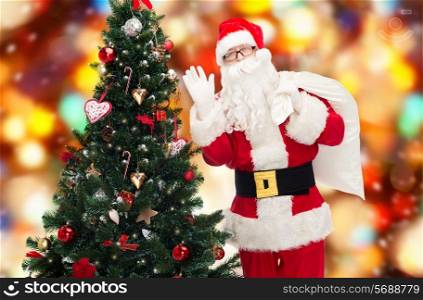 christmas, holidays and people concept - man in costume of santa claus with bag and christmas tree waving hand over red lights background