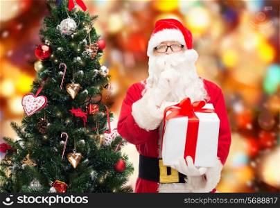 christmas, holidays and people concept - man in costume of santa claus with gift box and tree making hush gesture over red lights background