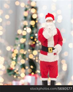 christmas, holidays and people concept - man in costume of santa claus making hush gesture over tree lights background