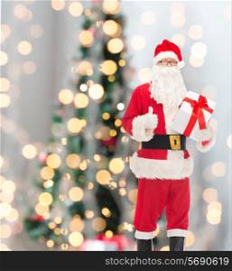 christmas, holidays and people concept - man in costume of santa claus with gift box showing thumbs up gesture over tree lights background