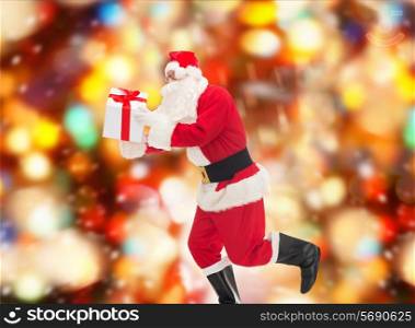 christmas, holidays and people concept - man in costume of santa claus running with gift box over red lights background