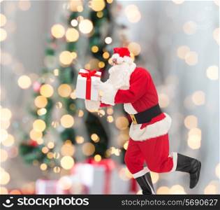 christmas, holidays and people concept - man in costume of santa claus running with gift box over tree lights background