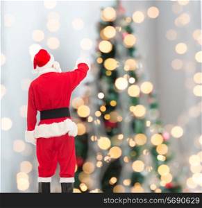 christmas, holidays and people concept - man in costume of santa claus pointing finger from back over tree lights background