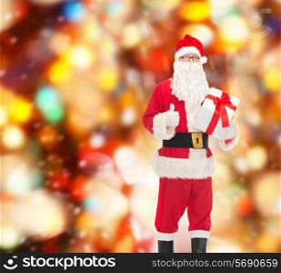 christmas, holidays and people concept - man in costume of santa claus with gift box showing thumbs up gesture over red lights background