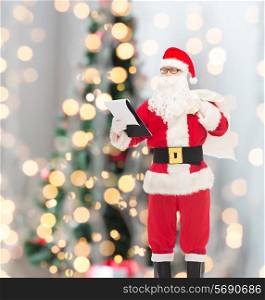 christmas, holidays and people concept - man in costume of santa claus with notepad and bag over tree lights background