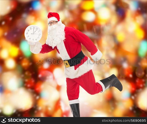 christmas, holidays and people concept - man in costume of santa claus running with clock showing twelve over red lights background