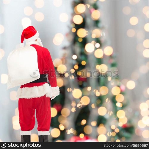 christmas, holidays and people concept - man in costume of santa claus with bag from back over tree lights background