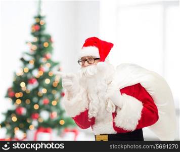 christmas, holidays and people concept - man in costume of santa claus with bag pointing finger over living room with tree