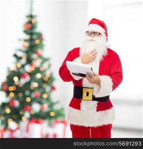 christmas, holidays and people concept - man in costume of santa claus with notepad over living room with tree background