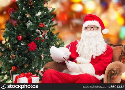 christmas, holidays and people concept - man in costume of santa claus with notepad, pen and christmas tree sitting in armchair over red lights background