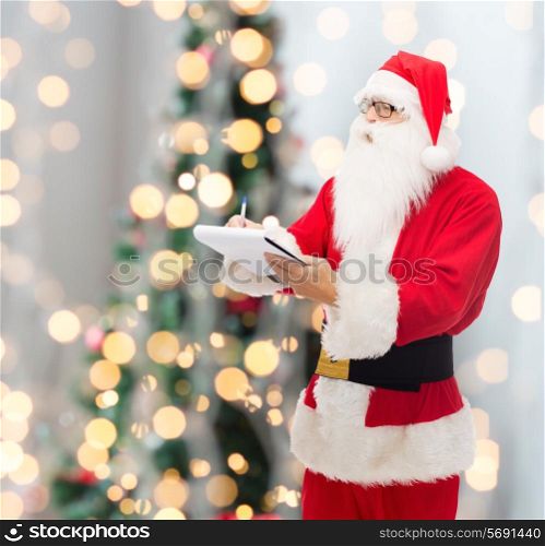 christmas, holidays and people concept - man in costume of santa claus with notepad and pen over tree lights background