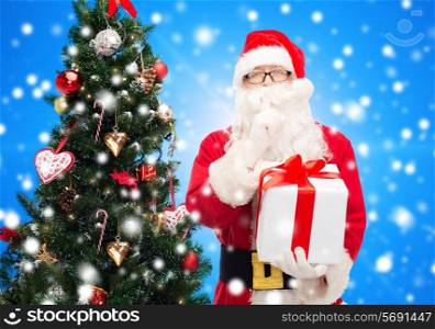 christmas, holidays and people concept - man in costume of santa claus with gift box and tree making hush gesture over blue lights background over blue snowy background