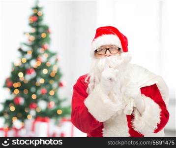 christmas, holidays and people concept - man in costume of santa claus with bag making hush gesture over living room with tree