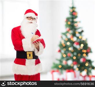 christmas, holidays and people concept - man in costume of santa claus with notepad and pen over living room with tree background