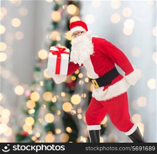 christmas, holidays and people concept - man in costume of santa claus running with gift box over tree lights background