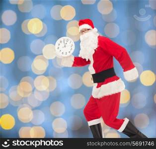 christmas, holidays and people concept - man in costume of santa claus running with clock showing twelve over blue lights background