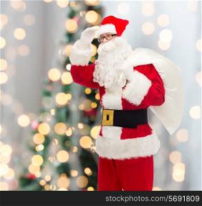 christmas, holidays and people concept - man in costume of santa claus with bag looking far away over tree lights background