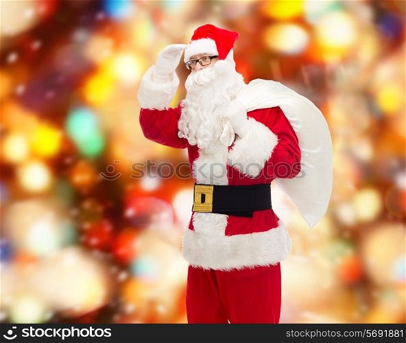 christmas, holidays and people concept - man in costume of santa claus with bag looking far away over red lights background