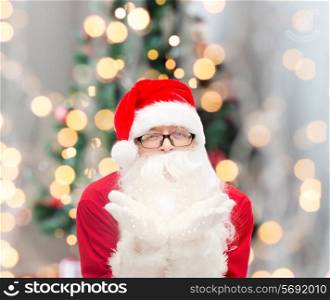 christmas, holidays and people concept - man in costume of santa claus blowing on palms over tree lights background