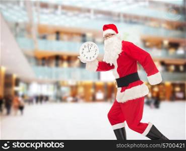 christmas, holidays and people concept - man in costume of santa claus running with clock showing twelve over shopping center background