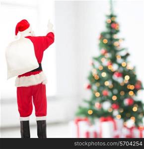 christmas, holidays and people concept - man in costume of santa claus with bag pointing finger from back over yellow lights background over living room and tree background