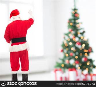 christmas, holidays and people concept - man in costume of santa claus writing something from back over living room and tree background