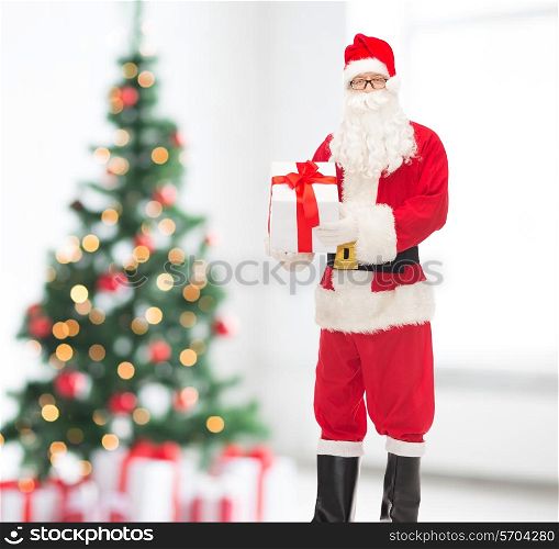 christmas, holidays and people concept - man in costume of santa claus with gift box over living room and tree background