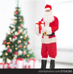 christmas, holidays and people concept - man in costume of santa claus with gift box over living room and tree background
