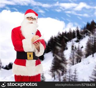 christmas, holidays and people concept - man in costume of santa claus with notepad and pen over snowy mountains background