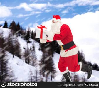 christmas, holidays and people concept - man in costume of santa claus running with gift box over snowy mountains background