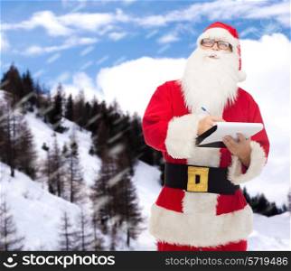 christmas, holidays and people concept - man in costume of santa claus with notepad and pen over snowy mountains background
