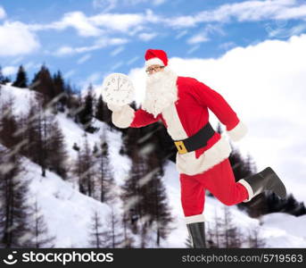 christmas, holidays and people concept - man in costume of santa claus running with clock showing twelve over snowy mountains background