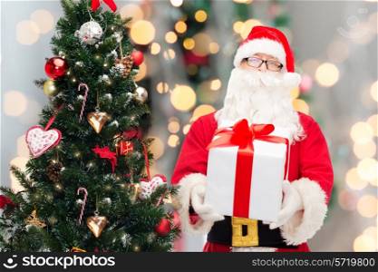 christmas, holidays and people concept - man in costume of santa claus with gift box and tree over lights background
